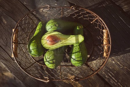 Photo for Top view of ripe whole and halved avocado in metal basket placed on wooden table on sunny day - Royalty Free Image