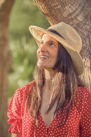 Photo for Portrait of positive young female in stylish dress and straw hat in countryside - Royalty Free Image
