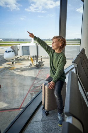Photo for Full body of excited little boy with curly blond hair in casual clothes sitting on suitcase near big panoramic window and pointing away while waiting for flight in modern airport - Royalty Free Image