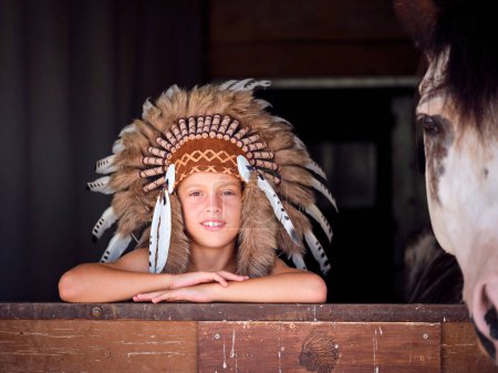 Téléchargez les photos : Enfant joyeux portant un authentique casque amérindien appuyé sur la barrière de l'écurie près du cheval de culture et regardant la caméra - en image libre de droit
