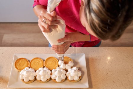 Téléchargez les photos : Vue du dessus de la culture pâtissière anonyme femme chef faisant des pics de crème sur les tartes à l'aide d'un sac de tuyauterie - en image libre de droit