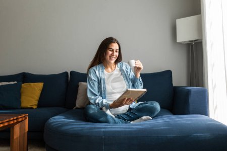 Photo for Positive pregnant female sitting on soft couch with notebook and drinking hot beverage - Royalty Free Image