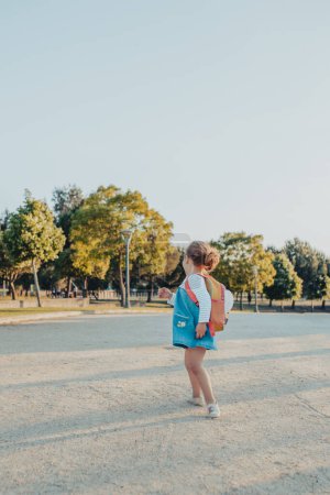 Photo for Side view cute little girl with backpack smiling while standing on sandy ground in park - Royalty Free Image