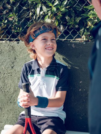 Téléchargez les photos : Souriant garçon préadolescent mignon en vêtements de sport assis avec raquette de tennis sur le banc près de grille métallique de clôture et levant les yeux tout en écoutant entraîneur - en image libre de droit