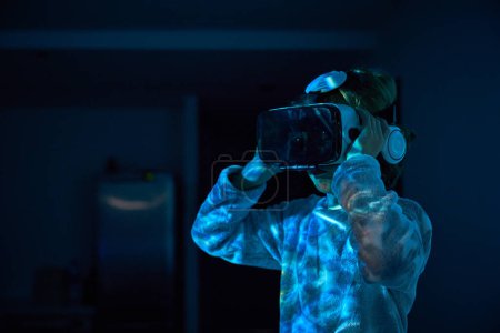 Téléchargez les photos : Petit garçon dans les écouteurs portant des lunettes VR et regardant la vidéo dans la pièce sombre avec la lumière du moniteur - en image libre de droit
