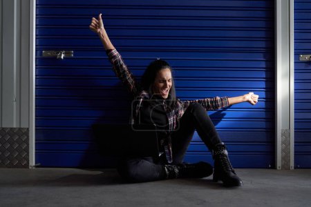 Photo for Full body of delighted female in casual wear showing thumb up gesture while sitting near metal blue gate of self storage - Royalty Free Image