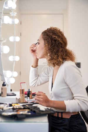 Téléchargez les photos : Vue latérale de la jeune femme aux cheveux bouclés faisant maquillage des yeux avec des produits cosmétiques assis devant un miroir éclairé - en image libre de droit