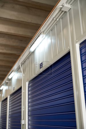 Photo for From below of numbered storage items with blue metal roller doors and bright lamps in modern storehouse - Royalty Free Image