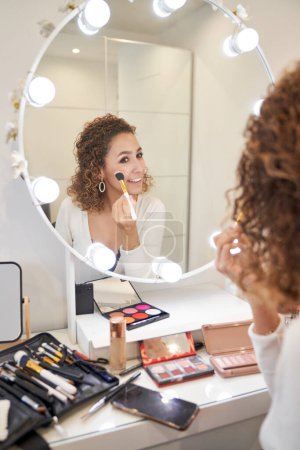 Foto de Reflejo espejo de una joven sonriente maquillando mejillas con cepillo profesional y rubor - Imagen libre de derechos