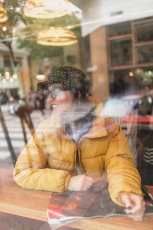 Téléchargez les photos : Vue latérale de la jeune femme attrayante dans le magazine de lecture de tenue élégant et regardant loin tout en étant assis derrière la fenêtre en verre dans le café - en image libre de droit