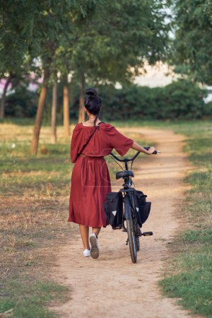 Téléchargez les photos : Vue arrière d'une femme méconnaissable en robe maxi rouge se promenant le long d'arbres verts sur un sentier étroit avec vélo près d'une prairie herbeuse - en image libre de droit