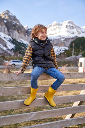 Foto de Cuerpo completo de niño sonriente con ropa casual y botas de goma sentado en una valla de madera mientras mira hacia otro lado en la zona montañosa - Imagen libre de derechos
