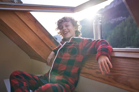 Téléchargez les photos : Enfant aux cheveux blonds souriants en pyjama à carreaux rouges assis près d'une grande fenêtre ouverte s'appuyant sur le rebord de la fenêtre le matin dans un hôtel situé près des montagnes - en image libre de droit