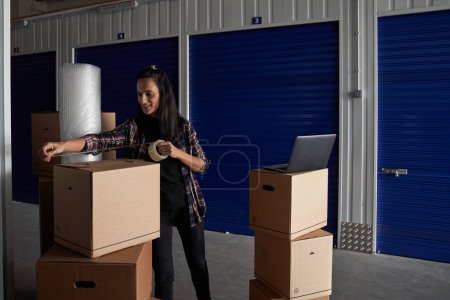 Photo for Content female using scotch to tape carton box with personal belongings near modern laptop before renting self storage during moving day - Royalty Free Image