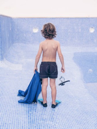 Téléchargez les photos : Vue arrière du corps complet torse nu préadolescent garçon dans les nageoires et avec serviette et masque de plongée debout sur le fond de la piscine vide - en image libre de droit