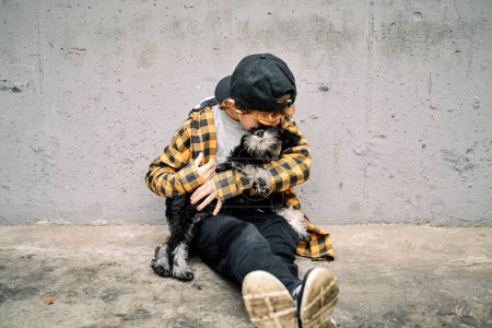 Photo for Full body of anonymous kid sitting against concrete wall and embracing little purebred dog while spending time together - Royalty Free Image