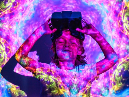 Téléchargez les photos : Enfant ravi avec les yeux fermés avec des lunettes VR sur la tête après avoir joué dans le monde virtuel tout en se tenant sous un éclairage abstrait coloré lumineux projecteur - en image libre de droit