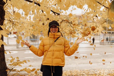 Foto de Hermosa joven en ropa de abrigo elegante sonriendo y tocando hojas amarillas en el árbol en increíble día de otoño en la calle de la ciudad - Imagen libre de derechos