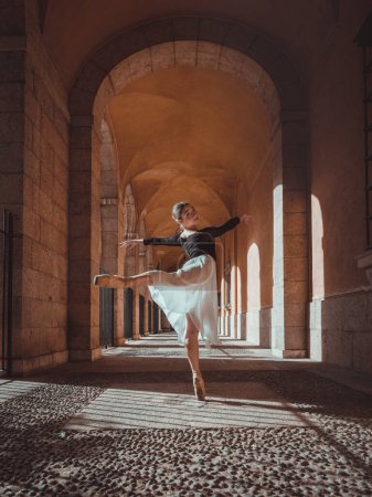 Téléchargez les photos : Vue latérale du corps entier d'une jeune danseuse de ballet gracieuse exécutant une position arabesque dans un couloir voûté - en image libre de droit