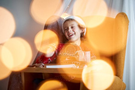 Téléchargez les photos : Garçon souriant dans le chapeau de Père Noël écrivant une lettre au plateau de lit avec du cacao et des biscuits dans la pièce lumineuse avec bokeh pendant les vacances de Noël - en image libre de droit