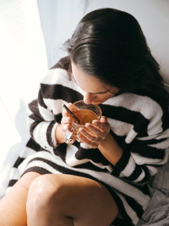 Foto de Desde arriba de la mujer sin rostro en suéter a rayas beber té caliente de la taza de vidrio mientras descansa en la cama cerca de la ventana en casa - Imagen libre de derechos