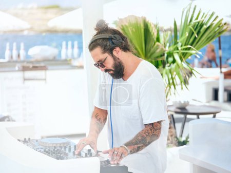 Foto de Vista lateral de DJ macho tatuado barbudo en camiseta blanca y auriculares ajustando el sonido de la música mientras está de pie en el controlador en la zona de fiesta del hotel resort con bahía en el fondo - Imagen libre de derechos