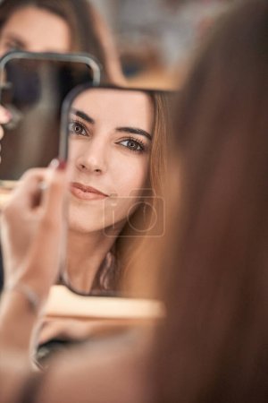 Téléchargez les photos : Crop content modèle féminin avec maquillage et cheveux longs regardant miroir à la main tout en étant assis dans un salon de beauté pendant le rendez-vous - en image libre de droit
