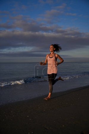 Photo for Full body of optimistic barefoot Hispanic female running on sandy beach along sea during workout in nature on evening time - Royalty Free Image