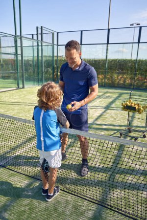 Photo for Full body of trainer speaking with boy in activewear while standing near net during padel tennis training on court in summer day - Royalty Free Image