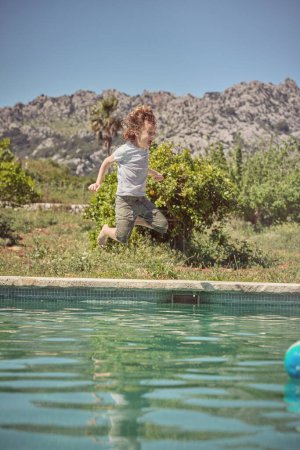 Téléchargez les photos : Plein corps vue latérale du garçon préadolescent heureux en vêtements décontractés sautant dans l'eau de la piscine et profitant de vacances d'été contre un paysage pittoresque - en image libre de droit