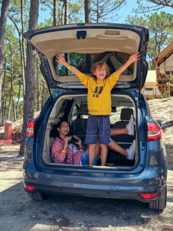 Photo for Full length of woman holding binoculars and looking at excited preteen boy standing in car trunk near glamping campsite in forest and enjoying freedom during summer vacation - Royalty Free Image