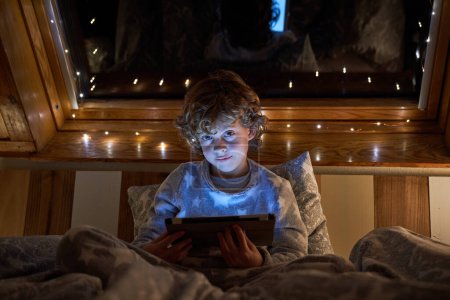 Foto de Contenido chico en ropa de dormir tableta de navegación y mirando a la cámara mientras está acostado en la cama debajo de la manta cerca de la ventana en la noche - Imagen libre de derechos