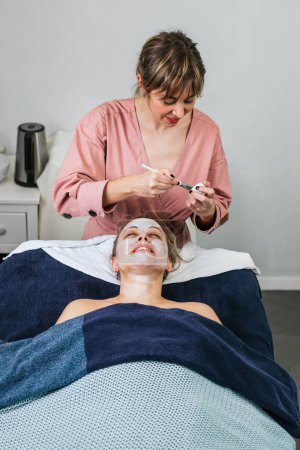 Foto de Cosmetólogo aplicando mascarilla facial hidratante en la cara del cliente femenino durante el procedimiento de cuidado de la piel en el salón de belleza moderno - Imagen libre de derechos
