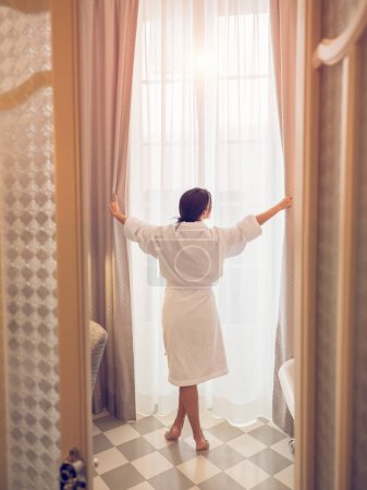 Téléchargez les photos : Vue arrière de la femme anonyme pieds nus en peignoir debout près de la fenêtre et ouvrant les rideaux dans la salle de bain de l'hôtel après le réveil le matin - en image libre de droit