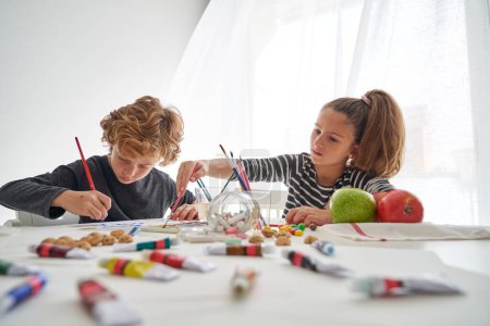 Téléchargez les photos : Enfants focalisés peignant avec des peintures colorées tout en étant assis ensemble à la table blanche salissante avec des fournitures d'art dispersées dans la salle de lumière - en image libre de droit
