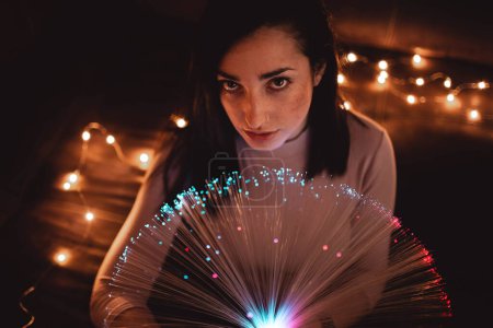 Photo for From above attractive brunette lady holding LED luminaire with fiber optic lights between illuminated fairy lights in obscurity - Royalty Free Image