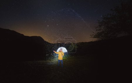 Téléchargez les photos : Voyageur méconnaissable veste jaune debout seul parmi les arbres sombres avec bras levé et parapluie la nuit pluvieuse - en image libre de droit