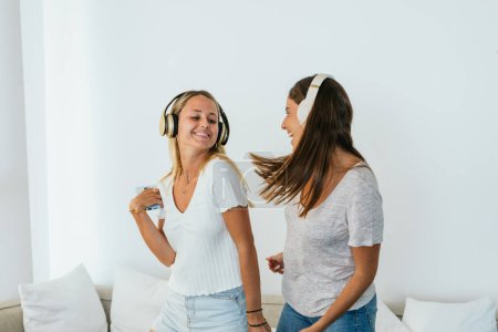 Foto de Amigas positivas escuchando música en auriculares inalámbricos mientras bailan y se divierten en la sala de estar ligera en casa - Imagen libre de derechos