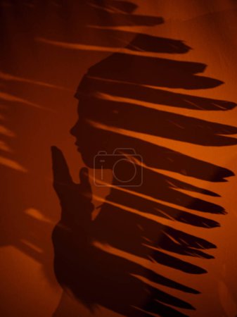 Photo for Side view silhouette on textile of unrecognizable boy in authentic Indian headdress standing with hand near mouth in dark room - Royalty Free Image
