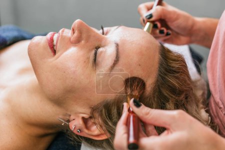 Téléchargez les photos : Crop maquilleuse anonyme appliquant de la poudre sur le visage du client féminin avec des brosses dans le centre de beauté - en image libre de droit