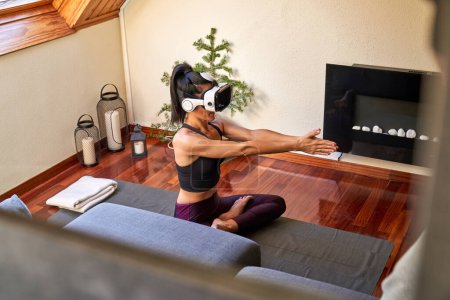 Téléchargez les photos : D'en haut femme mince en vêtements de sport en VR casque assis avec les jambes croisées et levé les bras droits tout en exerçant des poses de yoga à la maison - en image libre de droit