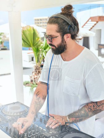 Téléchargez les photos : Disque jockey tatoué barbu concentré travaillant sur mixeur DJ tout en se tenant dans la zone de fête en plein air à la terrasse d'été de l'hôtel station balnéaire - en image libre de droit