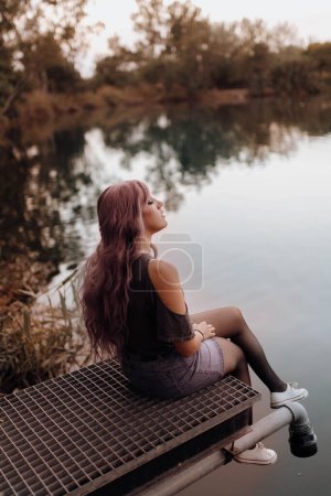 Foto de Vista lateral de la sensual mujer con el pelo rosa sentado en el muelle sobre el lago en paz - Imagen libre de derechos