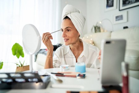 Foto de Vista lateral de una bloguera adulta concentrada con toalla en la cabeza aplicando sombra de cejas con pincel frente al espejo mientras filma un video de belleza en un teléfono inteligente - Imagen libre de derechos