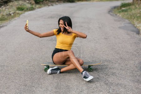 Foto de Hembra étnica tomando selfie en el teléfono inteligente mientras está sentado longboard en camino de asfalto - Imagen libre de derechos