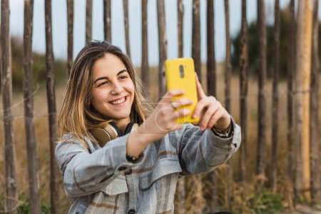 Téléchargez les photos : Jeune femme millénaire en vêtements décontractés et casque accroché sur le cou assis sur la promenade et prendre selfie sur smartphone dans la campagne - en image libre de droit