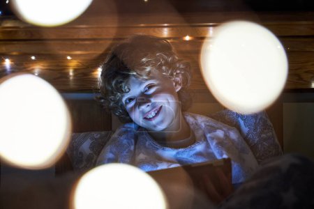 Téléchargez les photos : Garçon heureux en vêtements de nuit jouer au jeu sur tablette et en regardant la caméra tout en étant assis dans la chambre avec bokeh sur la nuit - en image libre de droit