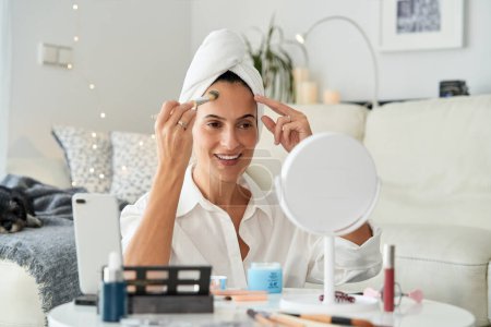 Téléchargez les photos : Femme adulte souriante positive avec serviette sur la tête appliquer le fond de teint sur le front avec brosse devant le miroir tout en tirant blog de beauté sur téléphone mobile à la maison - en image libre de droit