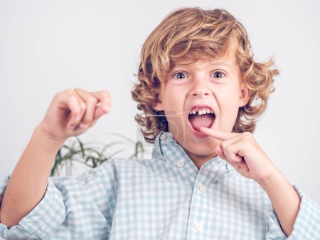 Téléchargez les photos : Adorable bouclé chevelu preteen garçon secousses dent avec fil et processus de démonstration avec bouche ouverte et en regardant la caméra - en image libre de droit