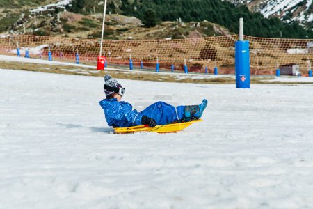 Foto de Vista lateral del niño en ropa de abrigo a caballo trineo de plástico en la nieve cerca de la cresta de montaña en el día de fin de semana en la estación de esquí - Imagen libre de derechos